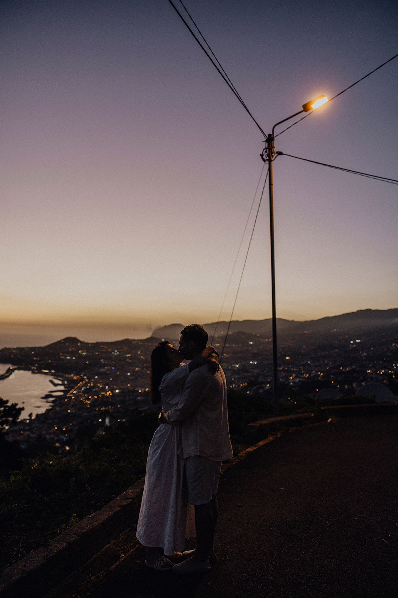 K&D obejmujący się pod latarnią uliczną w Funchal, Madera, z widokiem na rozświetlone miasto i ocean o zmierzchu, magiczna godzina, sesja poślubna by Camera Folks Wedding Photo & Video.