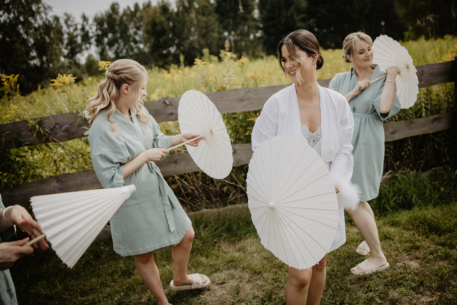 Panna Młoda K z dwiema druhnami, bawiące się białymi parasolkami, na tle zieleni i drewnianego płotu w Sielsko Anielsko, sfotografowane przez Camera Folks – Wedding Photo & Video.