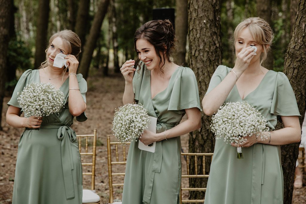Trzy druhny w zielonych sukniach, ocierające łzy wzruszenia, trzymające bukiety z gipsówki, podczas ceremonii ślubnej w Sielsko Anielsko, sfotografowane przez Camera Folks – Wedding Photo & Video.