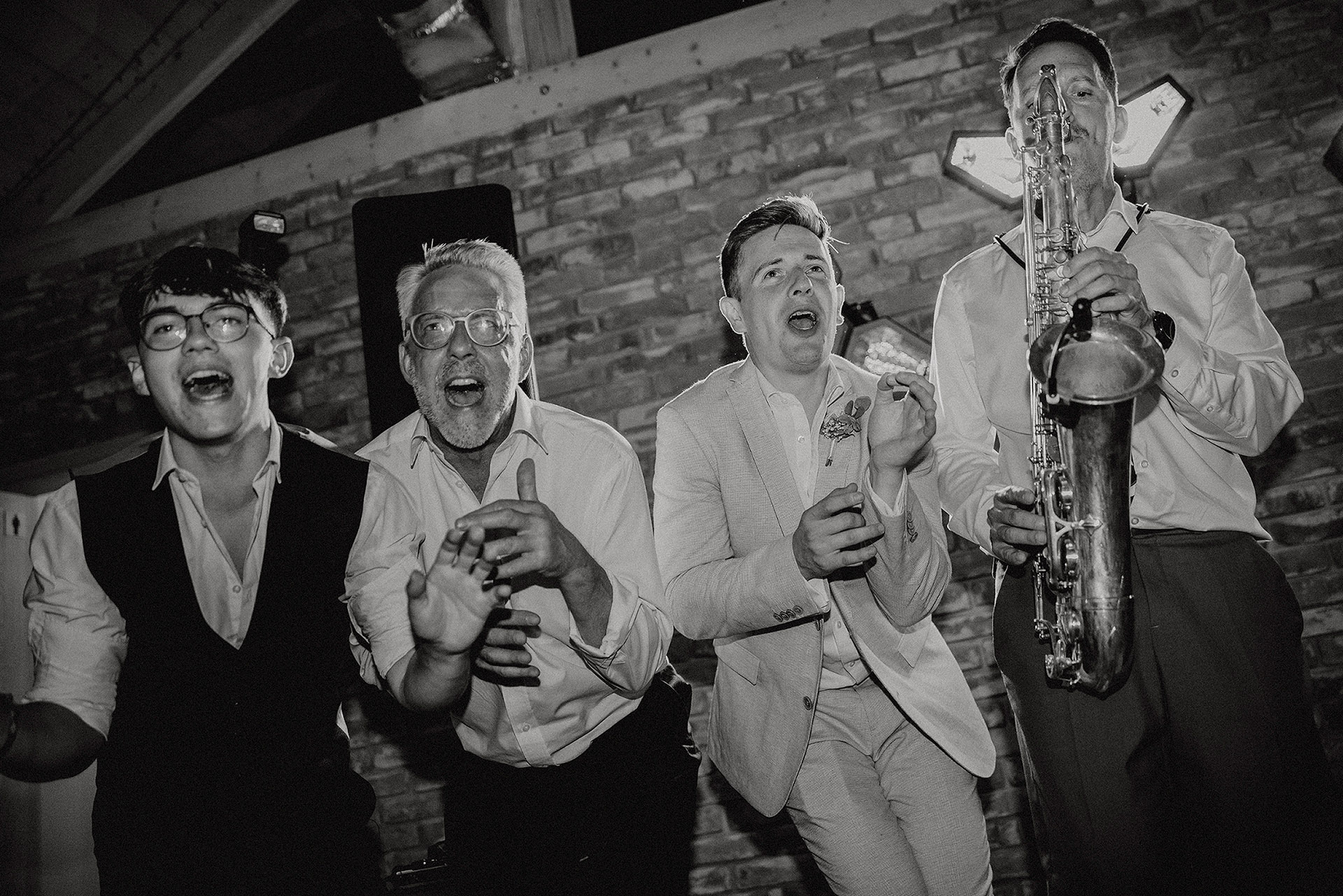 Czarno-białe zdjęcie saksofonisty (@weddandsax) grającego na saksofonie, otoczonego rozśpiewanymi i rozbawionymi mężczyznami (w tym Świadek i Dziadek Panny Młodej z poprzednich zdjęć), wszyscy śpiewający lub gestykulujący, podczas wesela w Sielsko Anielsko, sfotografowane przez Camera Folks – Wedding Photo & Video.