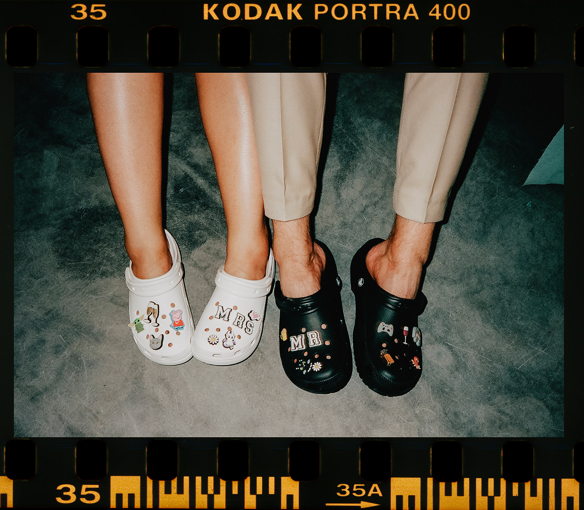 Skan z kliszy Kodak Portra 400 przedstawiający stopy Panny Młodej w białych Crocsach z napisem "Mrs." i Pana Młodego w czarnych Crocsach z napisem "Mr.", ozdobionych zabawnymi przypinkami, symbolizujące zmianę obuwia po weselu, sfotografowane przez Camera Folks – Wedding Photo & Video.
